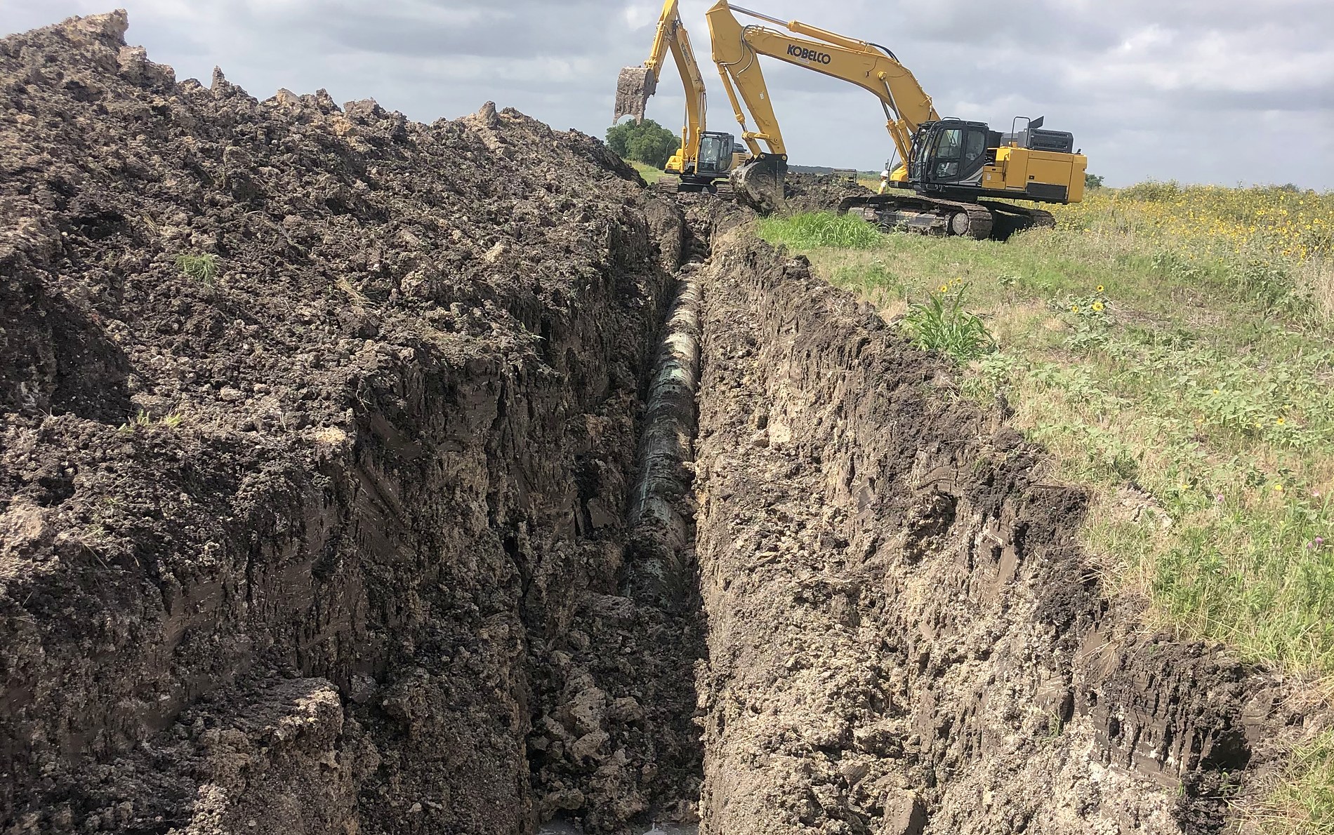 Open trench with exposed line pipe and an excavator working the right-of-way